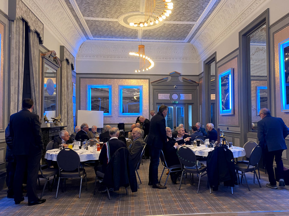 Photograph of a large high-ceilinged room with grey walls and white ceiling. Filling the room are four round tables that each seat 8 people. Most people are seated with drinks and cutlery set out ready for a meal, while a few others are standing and talking, or making their way to their seats.