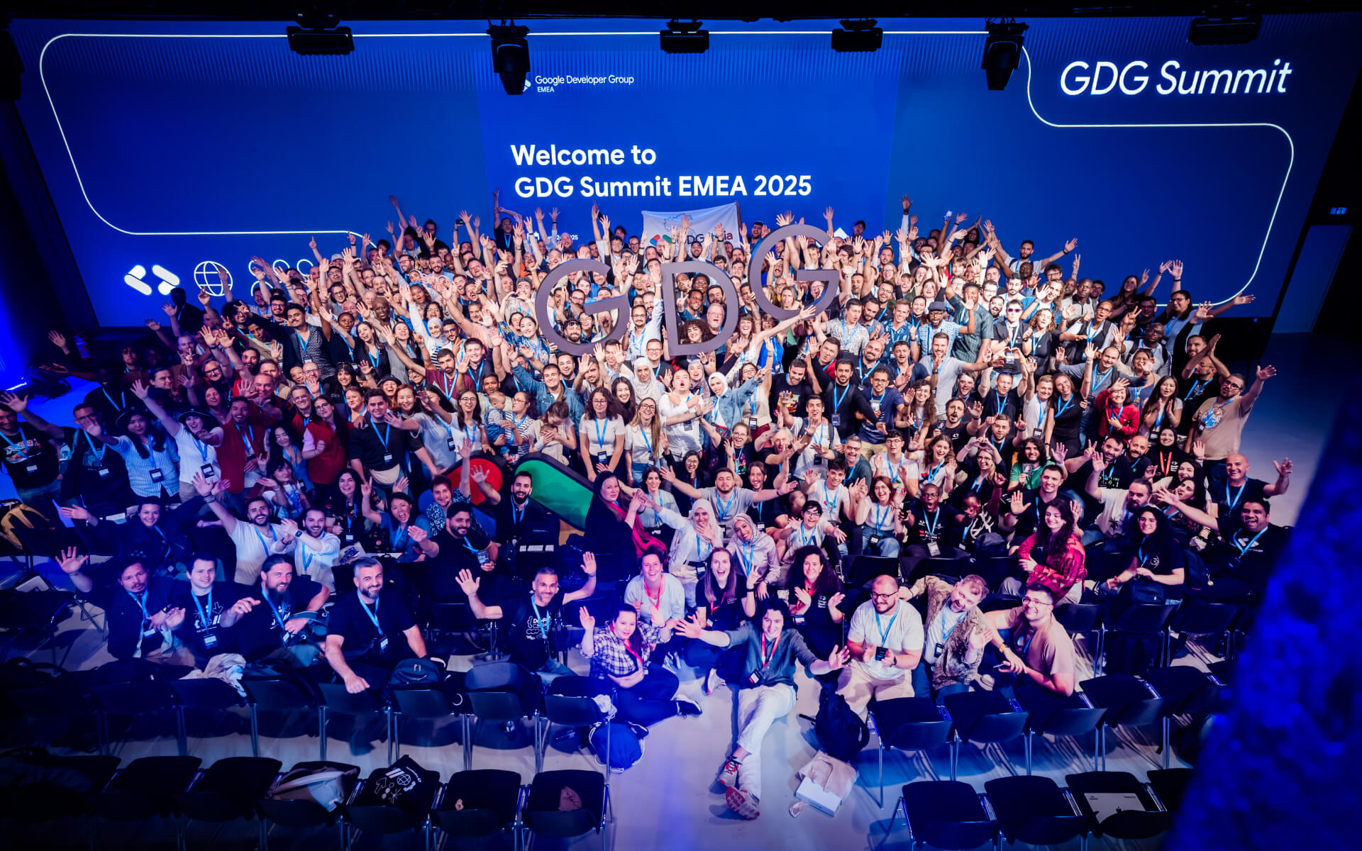 A group photograph taken looking down at hundreds of people with their hands in the air, some holding large letters that spell out GDG. They are in front of a backdrop that reads 'Welcome to GDG Summit EMEA 2025'.