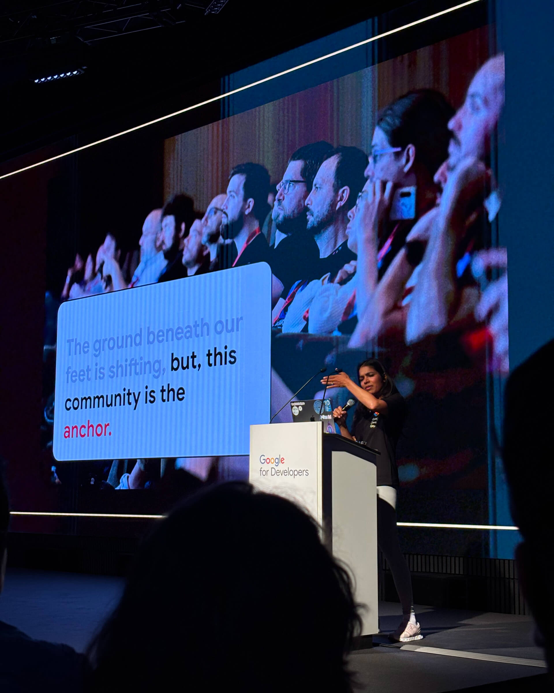 Kamal Sandu stands behind a podium that says 'Google for Developers'. She is holding a microphone and gesturing towards a large presentation slide beside her that reads 'The ground beneath our feet is shifting, but, this community is the anchor'.