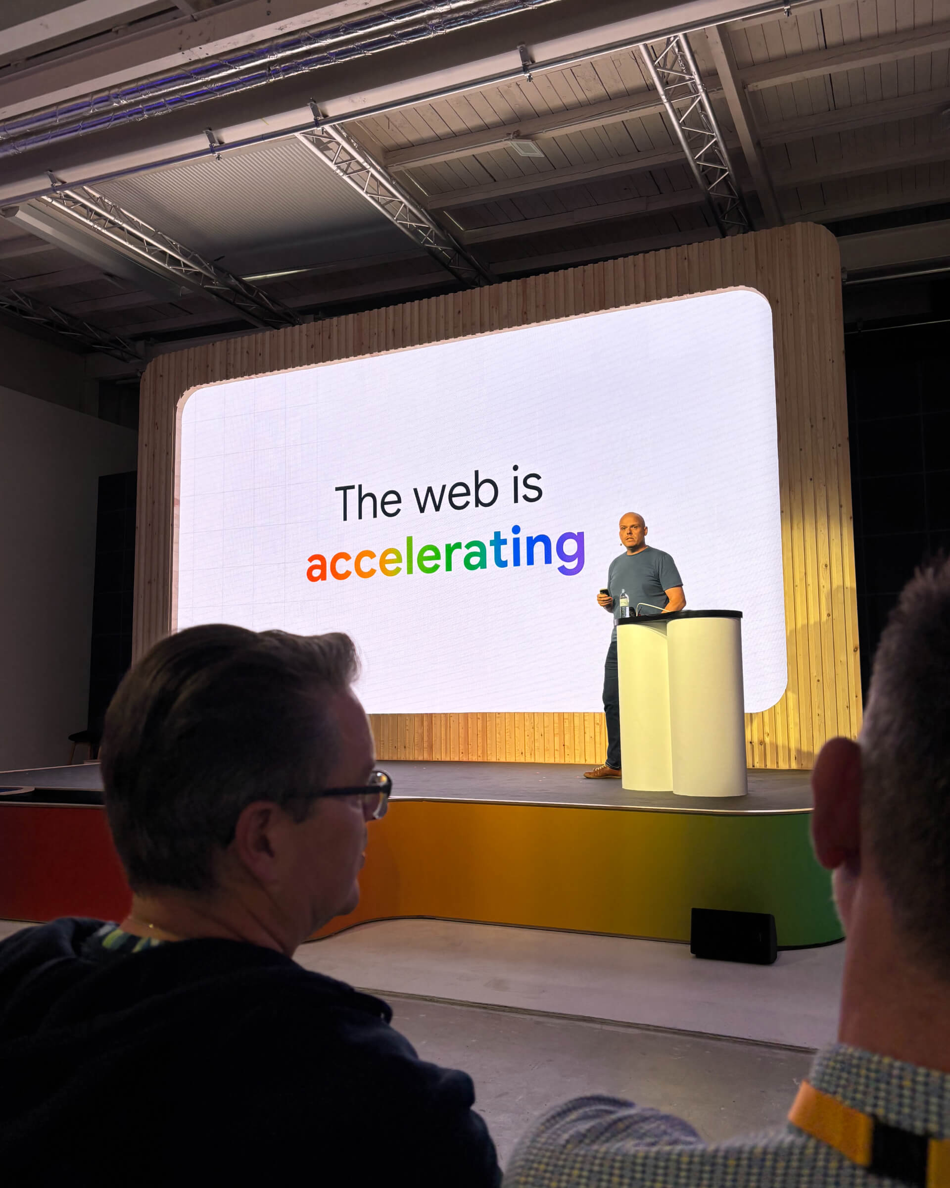 Paul Kinlan stands on a stage holding a presentation clicker in front of a large slide that reads 'The web is accelerating'.