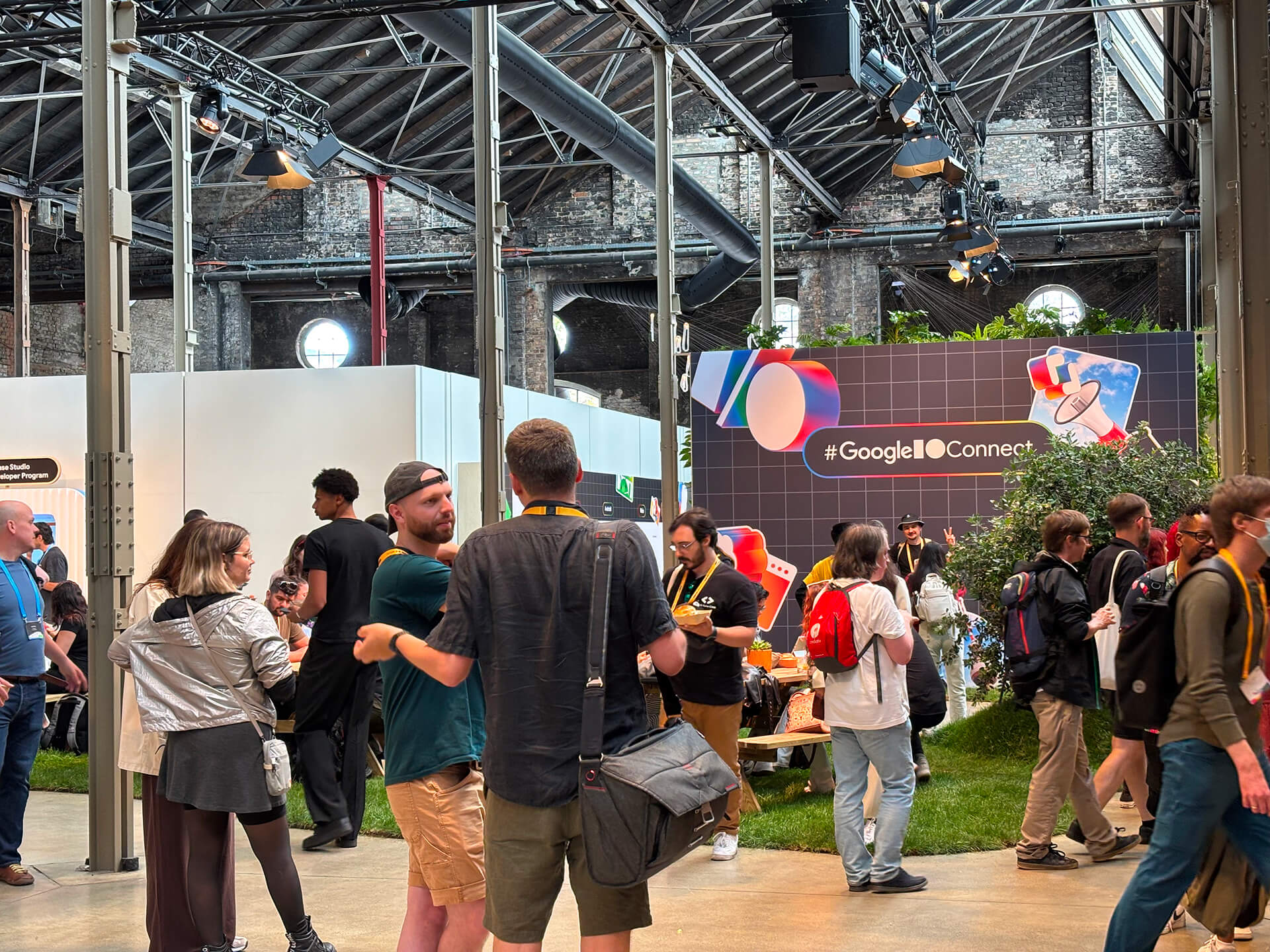 A large warehouse with high ceilings and exposed bricks, filled with people standing while talking. A sign in the background reads '#GoogleIOConnect'. There are areas of grass turf and plants in the background.