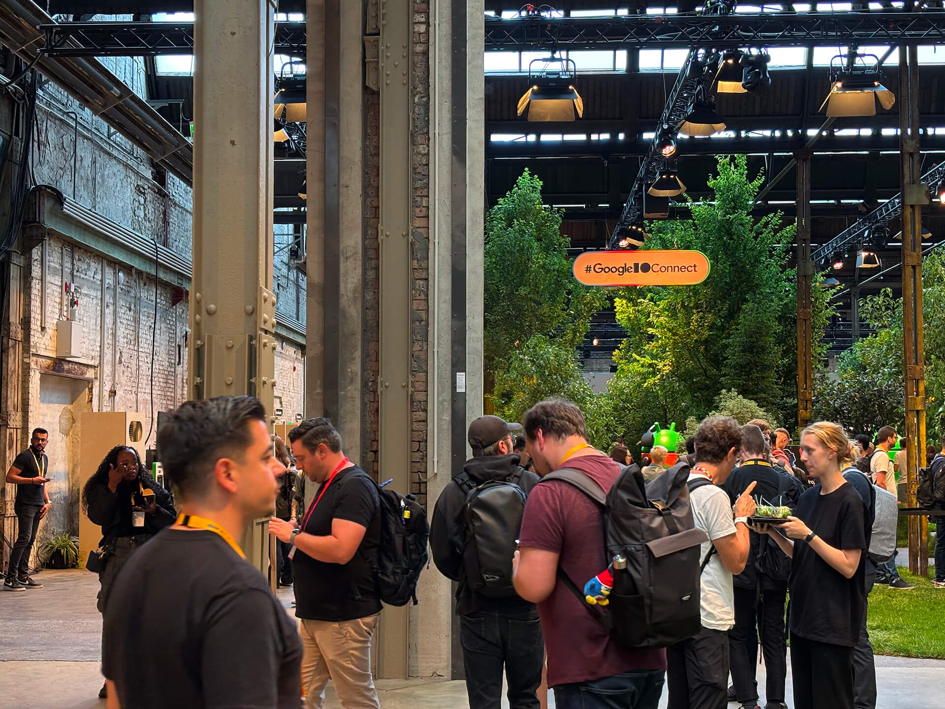 A large warehouse with high ceilings and exposed bricks, filled with people standing while talking and eating small bowls of food. An orange sign above reads '#GoogleIOConnect'. In the background there is a large indoor garden with trees, bushes and grass floor.