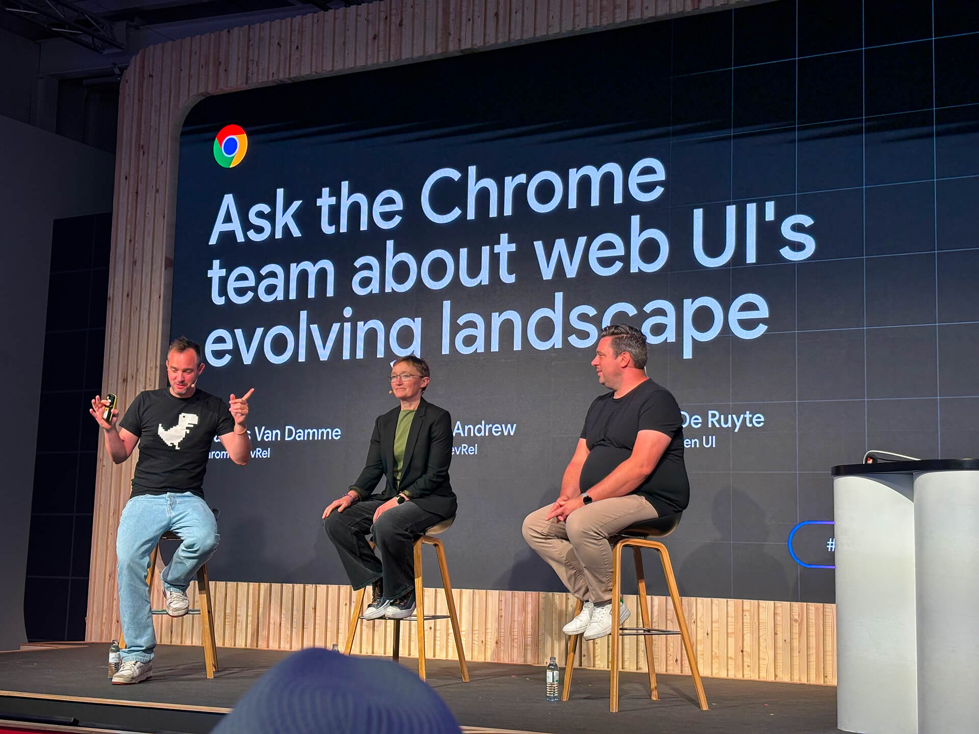 Bramus Van Damme, Rachel Andrew, and Brecht de Ruyte sat on high stools on a stage in front of a large presentation slide that reads 'Ask the Chrome team about web UI's evolving landscape'. Bramus is gesturing his hands outwards as he speaks.