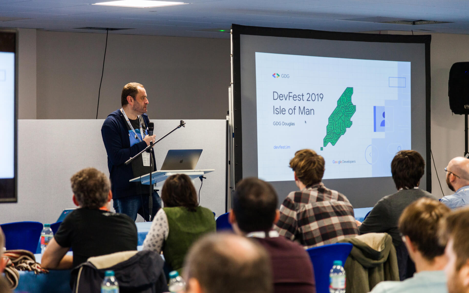John Middleton stood on a stage in front of a room full of people. He is holding a microphone and stood next to a large slide projected onto the wall that reads 'DevFest 2019 Isle of Man'.