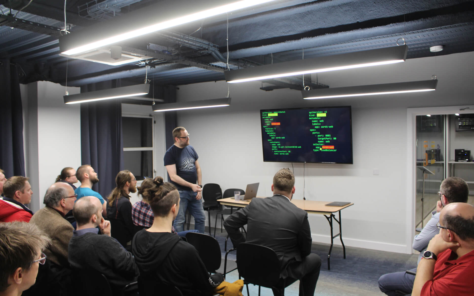 John Middleton stood in front of a room of people looking a large screen of code as he presses a presentation clicker to change slides.